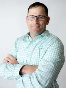 Man with short, black hair wearing glasses and a green plaid shirt is crossing his arms posing for his headshot against a white and gray background